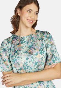 Kurzärmeliges Kleid aus seidig glänzendem Stoff, mit grünem Grundton und mehrfarbigem Blumenmuster sowie Rundhalsausschnitt. Sanfte Textur, leicht.