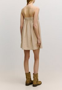 Lichtbeige jurk tot de knie met een smock achterkant, dunne bandjes en een relaxed fit, gecombineerd met bruin suède laarzen tot de enkels.