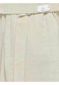 Ljust beige strukturerad tyg med ett korsmönster, som har en midja med en vit knapp och sidofält för extra designdetaljer.