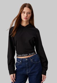 Čierna skrátená mikina s kapucňou má textúrovaný povrch, obsahuje šnúrku na kapucni a biely logo Calvin Klein Jeans na páse. Tmavomodré džíny.