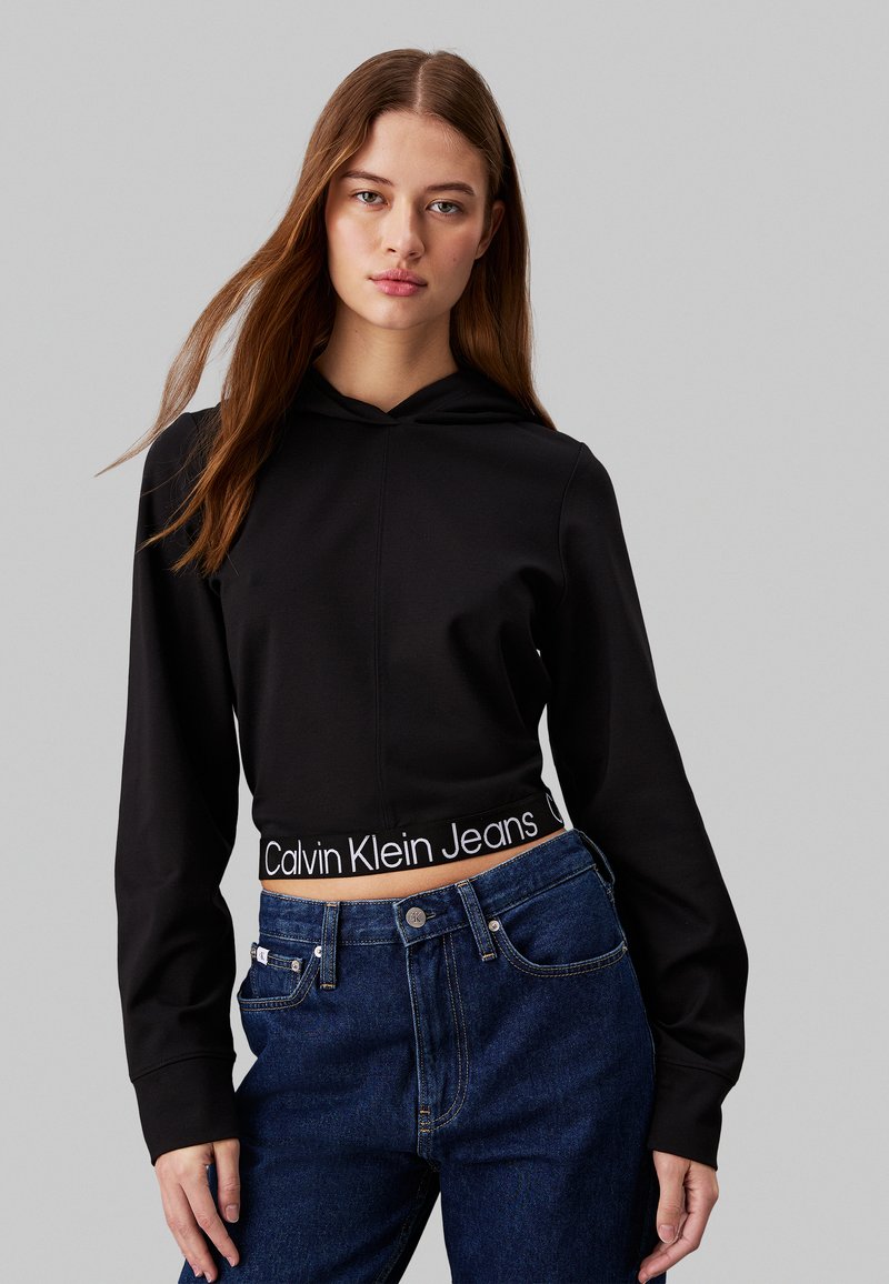 Čierna skrátená mikina s kapucňou má textúrovaný povrch, obsahuje šnúrku na kapucni a biely logo Calvin Klein Jeans na páse. Tmavomodré džíny.