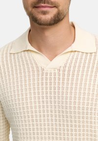 Sweter polo z dzianiny w kolorze kremowym, z dekoltem w serek, charakteryzujący się teksturowanym wzorem z małymi prześwitami. Kołnierzyk poprawia wentylację i styl.