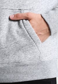 Felpa grigio chiaro con una texture morbida, dotata di una tasca frontale e orlo a coste. Mano parzialmente dentro la tasca.