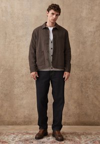 Young man wearing a brown buttoned jacket, grey cardigan, white shirt, dark jeans, and brown boots standing on a patterned rug.