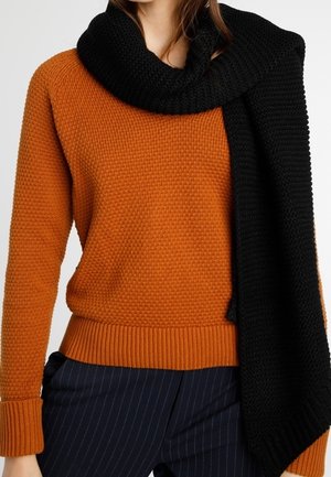 Femme portant un pull orange texturé, une écharpe noire posée sur l'épaule et un pantalon noir à fines rayures, vue du cou jusqu'aux hanches.