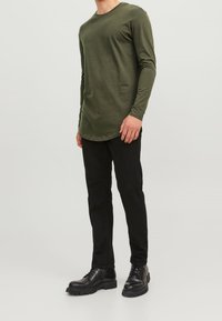 Olivi vihreä pitkähihaisessa paidassa on pyöristetty helma, yhdistettynä mustiin slim fit -housuihin ja mustiin nauhakengiin. Liu'uttava kangas.