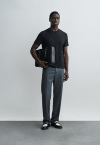 Homme en t-shirt noir, pantalon gris, mocassins noirs et blancs tenant un sac d'ordinateur portable noir, debout contre un mur gris uni.