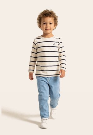 Bambino piccolo con capelli ricci che cammina, indossa una maglietta a maniche lunghe color crema con strisce nere, jeans azzurri chiari e scarpe da ginnastica bianche.