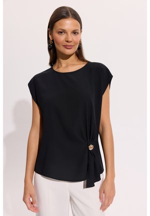 Kobieta ubrana w czarny top bez rękawów z złotym zapięciem po lewej stronie i białe spodnie, stojąca na jasnym tle.