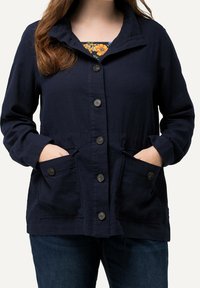 Kvinna som bär en mörkblå knappad jacka med stora framfickor och en blommig topp under, händerna instuckna i fickorna.