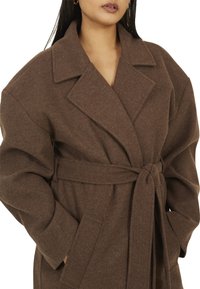 Cappotto in lana marrone con design avvolgente, vestibilità oversize, ampi risvolti, cintura da annodare in vita e tasche laterali. Tessuto morbido.