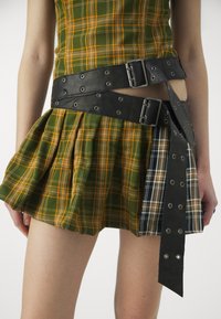 Svart läderbälte med metallhardware som omger en plisserad grön och gul rutiga kjol, med en kontrasterande rutig panel och texturerad tyg.