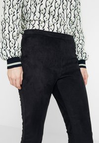 Czarne spodnie z zamszowego materiału o gładkiej fakturze, z wysokim stanem i dopasowanym krojem, w połączeniu z wzorzystą bluzką i paskami na mankietach.