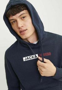Tengerészkék kapucnis pulcsi puha anyagból, fehér téglalap alakú logóval, piros felirattal: "JACK & JONES". Kenguru zseb és állítható kapucni.