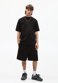Sort kortærmet t-shirt med en afslappet pasform, parret med sorte shorts. Materialet ser blødt ud. Tilbehør inkluderer en sølv kæderekæde.