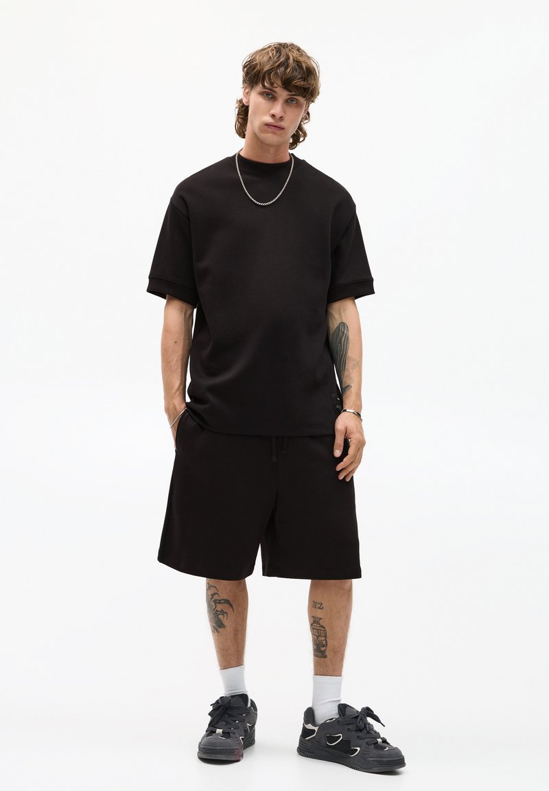 Sort kortærmet t-shirt med en afslappet pasform, parret med sorte shorts. Materialet ser blødt ud. Tilbehør inkluderer en sølv kæderekæde.