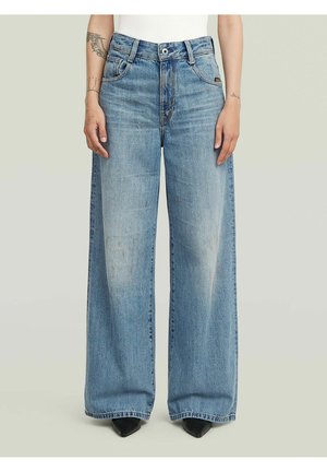 Une personne porte un jean large bleu clair avec une taille haute et des chaussures noires pointues, debout devant un fond uni gris clair.