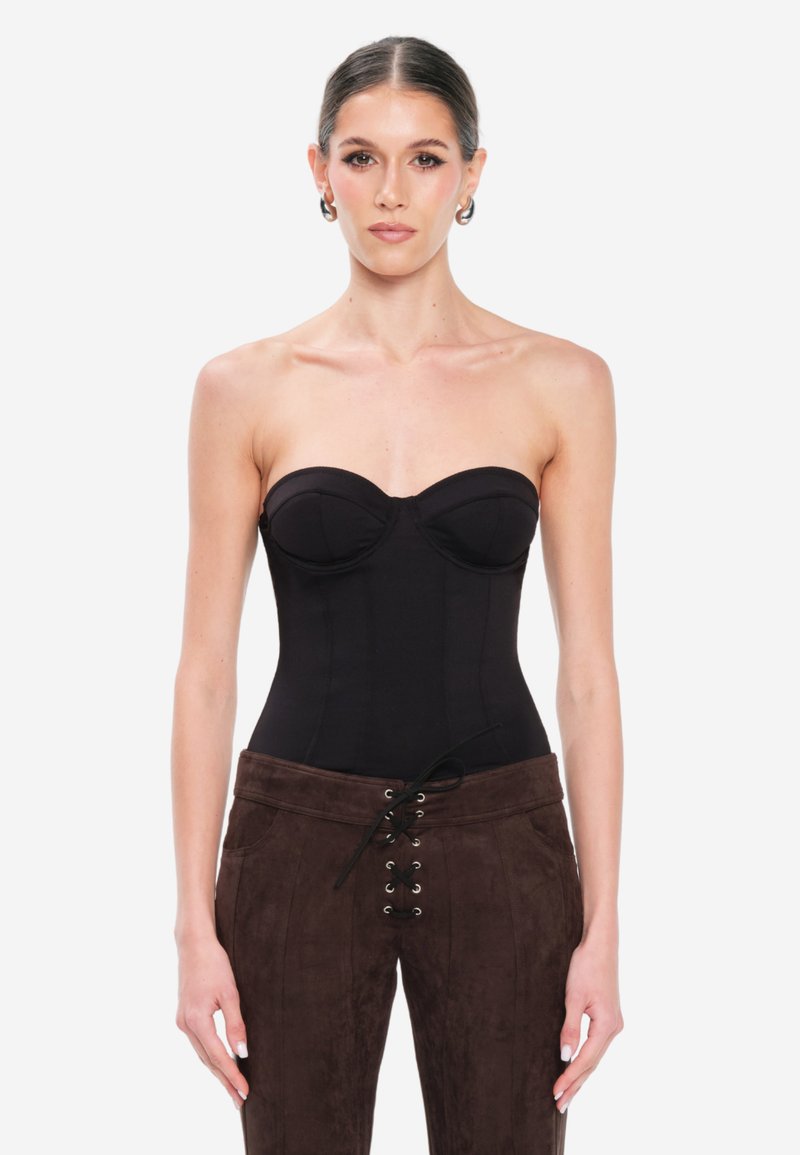 Donna con capelli raccolti, che indossa un corsetto nero senza spalline e pantaloni marroni con lacci, in piedi contro uno sfondo bianco semplice.
