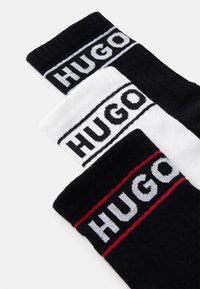 Tres pares de calcetines: negros con rayas blancas y rojas, con el nombre de la marca "HUGO" en letras grandes en las bandas superiores. Textura de tela suave.