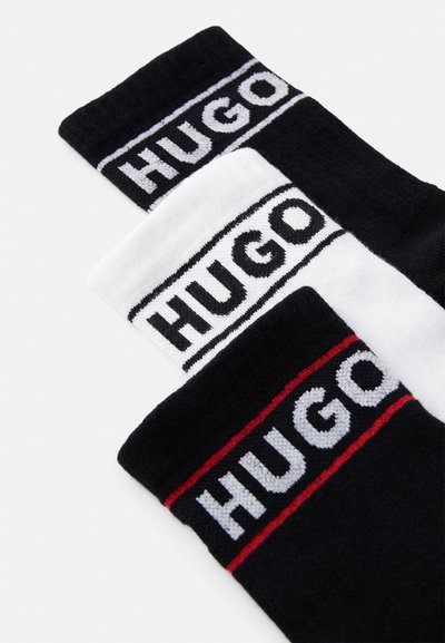 Trois paires de chaussettes : noires avec des rayures blanches et rouges, arborant le nom de la marque "HUGO" en lettres gras sur les bandes supérieures. Texture de tissu douce.