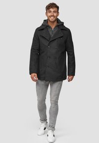 Een glimlachende man in een zwart dubbelzijdig jasje over een grijze hoodie, lichte grijze jeans en witte sneakers, die naar voren loopt op een grijze achtergrond.