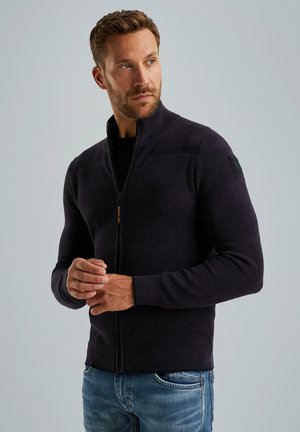 Tamno plavi zip-up jakna, teksturirana pletenina s horizontalnim prugama, visoki ovratnik i crna kvačica na ramenu. Nosiva s plavim trapericama.