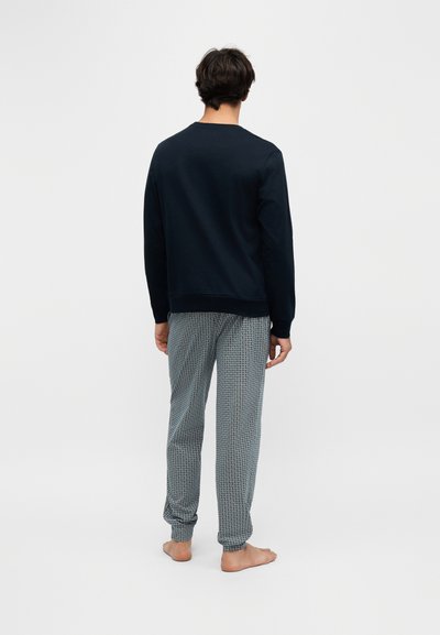 Homme debout pieds nus, de dos, portant un haut noir à manches longues et un pantalon de pyjama gris à motifs sur un fond uni.