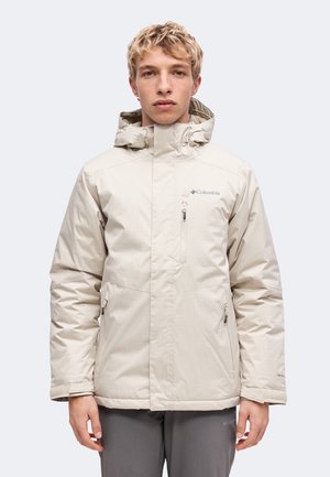 Ung person iført en lysebeige Columbia jakke med hætte, lynlåslommer og høj krave, stående foran en ensfarvet baggrund.