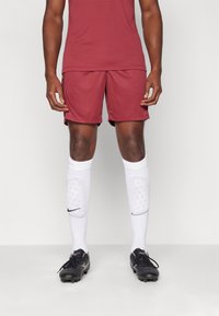 Camisa deportiva marrón y pantalones cortos combinados con calcetas blancas hasta la rodilla que presentan un logo negro, y botines de fútbol negros con superficie texturizada.