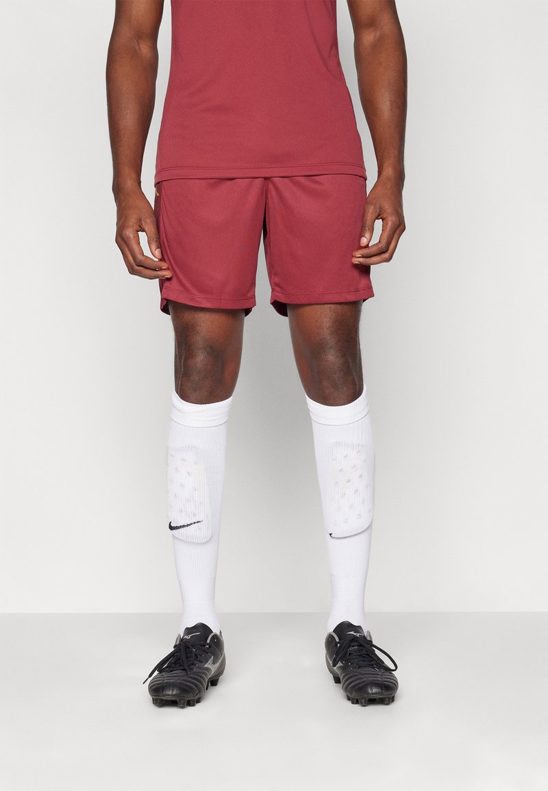 Camisa deportiva marrón y pantalones cortos combinados con calcetas blancas hasta la rodilla que presentan un logo negro, y botines de fútbol negros con superficie texturizada.