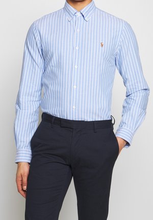 Mężczyzna w jasnoniebieskiej i białej koszuli w pionowe paski, zapiętej na guziki, w ciemnogranatowych spodniach, z ręką w kieszeni, stojący na jednolitym tle.