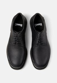 Un paio di scarpe nere in pelle con lacci, suole testurizzate e logo "CAMPER" visibile sulle solette interne, mostrate dall'alto su uno sfondo bianco.