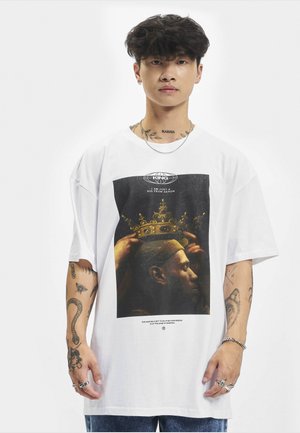 Junger Mann mit lockigem Haar und mehreren Tätowierungen, der ein weißes, übergroßes T-Shirt mit einer Krone und einem Porträt-Design trägt.