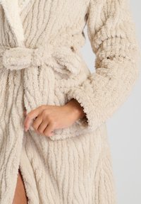 Beige pluche badjas met een gestructureerd, geribbeld ontwerp. Voorzien van een strikceintuur en brede mouwen. De oppervlakte is zacht en fluffy, wat het comfort vergroot.