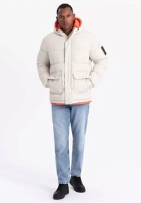 Puffig vit jacka med orange innanför, dragkedja, två framfickor och huva. Bärs över en orange skjorta med blå jeans och svarta skor.