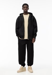 Črna zip-up kapuca z vlečnimi vrvicami, kombinirana z bež majico in črnimi trenirškimi hlačami. Obutev so svetlo bež superge. Preprosta zasnova.