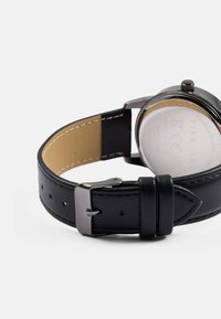 Czarny skórzany pasek do zegarka z beżowym wnętrzem, stalowa tylna część koperty, srebrne elementy sprzętowe oraz dopasowane detale w kolorze nici.
