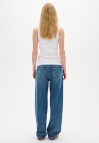 Femme aux longs cheveux blonds portant un haut blanc sans manches et un jean bleu à jambes larges, debout devant un fond blanc uni.