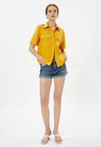 Camicia gialla a maniche corte con due tasche sul petto, abbinata a pantaloni in denim e ciabatte bianche. Tessuto morbido e vestibilità rilassata.