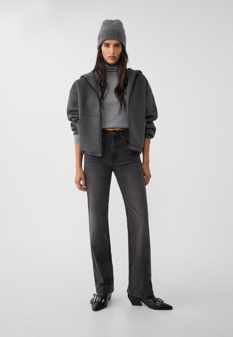 Hoodie grigio oversize, maglione a collo alto grigio attillato, jeans neri a vita alta e stivali neri con tacco a punta e dettagli decorativi in metallo.