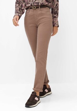 Vrouw draagt opgerolde bruine broek, paisley patroon blouse, zwarte riem en zwarte sneakers met roze accenten, lopend op witte achtergrond.