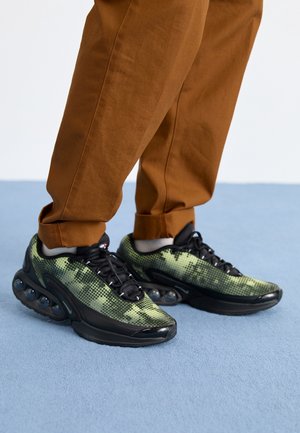 Persona con pantalones marrones y zapatillas con patrón pixelado verde y negro y amortiguación visible de aire, de pie sobre una alfombra azul claro.