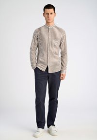 Camicetta a righe a maniche lunghe con colletto alla cinese in beige e marrone, abbinata a pantaloni blu navy e sneaker bianche. Texture liscia, design con bottoni.