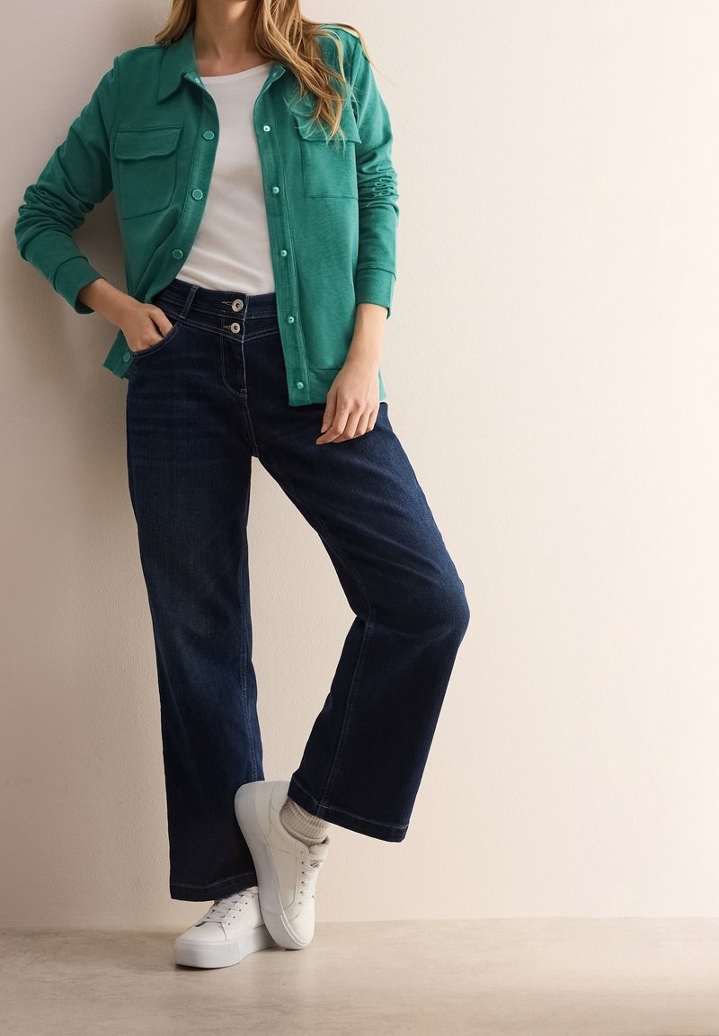 Femme portant une chemise boutonnée bleu sarcelle par-dessus un haut blanc, jean large foncé et baskets blanches, debout contre un mur beige.