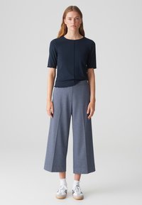 Haut bleu marine à manches courtes avec un ourlet festonné, associé à un pantalon large gris et des baskets grises avec des accents blancs. Fond uni.