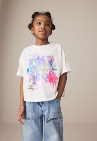 Hvid kortærmet t-shirt med farverige grafiske designs af blomster og en sommerfugl, kombineret med lyseblå baggy jeans.