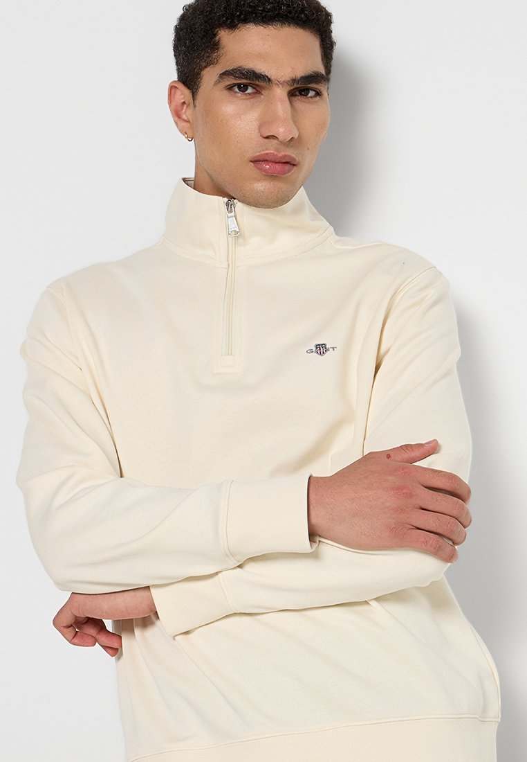 Ecrufarbenes Fleece-Pullover mit hohem Kragen, halbem Reißverschluss und gesticktem Logo auf der Brust. Weicher Stoff, lockere Passform.