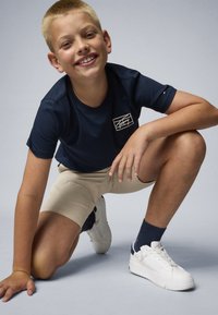 T-shirt bleu marine avec un petit logo blanc, short beige, baskets blanches et chaussettes bleu marine. Le garçon est à genoux avec une expression enjouée.