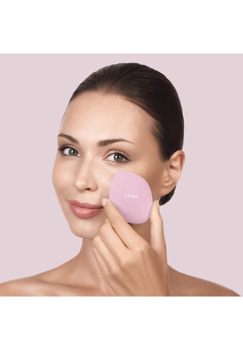 Woman with tied-back hair gently pressing a pink Geske facial device to her cheek against a pale purple background.