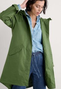 Veste légère verte avec capuche, fermeture à boutons-pression à l'avant et poches latérales, superposée à une chemise en denim bleu clair et un pantalon à cordon de serrage.